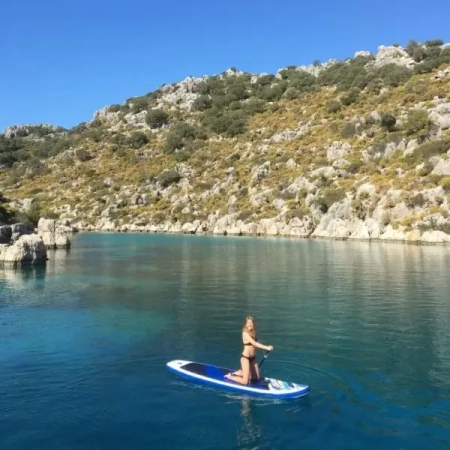 A woman paddleboarding on turquoise water during a Kas to Kekova and Back 3 Nights gulet cruise, surrounded by rocky hills and lush Mediterranean landscape.