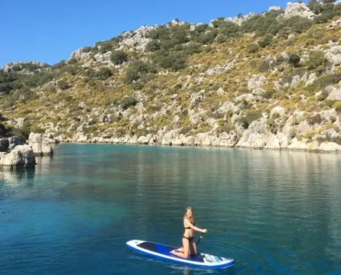 A woman paddleboarding on turquoise water during a Kas to Kekova and Back 3 Nights gulet cruise, surrounded by rocky hills and lush Mediterranean landscape.