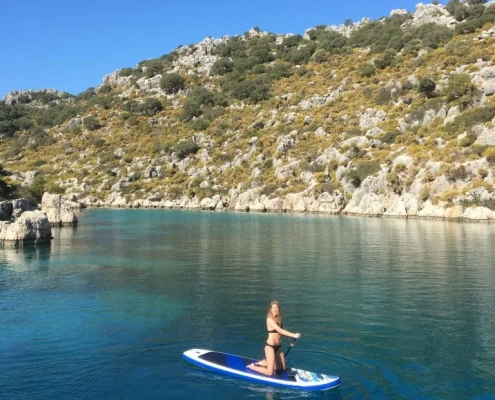 A woman paddleboarding on turquoise water during a Kas to Kekova and Back 3 Nights gulet cruise, surrounded by rocky hills and lush Mediterranean landscape.