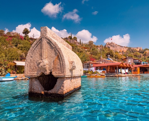 Ancient Lycian sarcophagus in the water at Kalekoy during the Demre to Kas and Back gulet cruise, with village houses and Simena Castle in the background