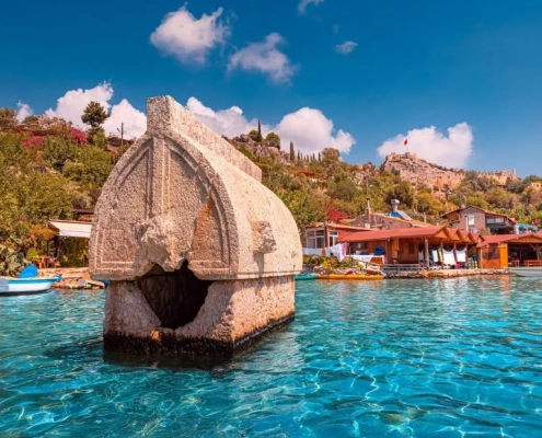 Ancient Lycian sarcophagus in the water at Kalekoy during the Demre to Kas and Back gulet cruise, with village houses and Simena Castle in the background