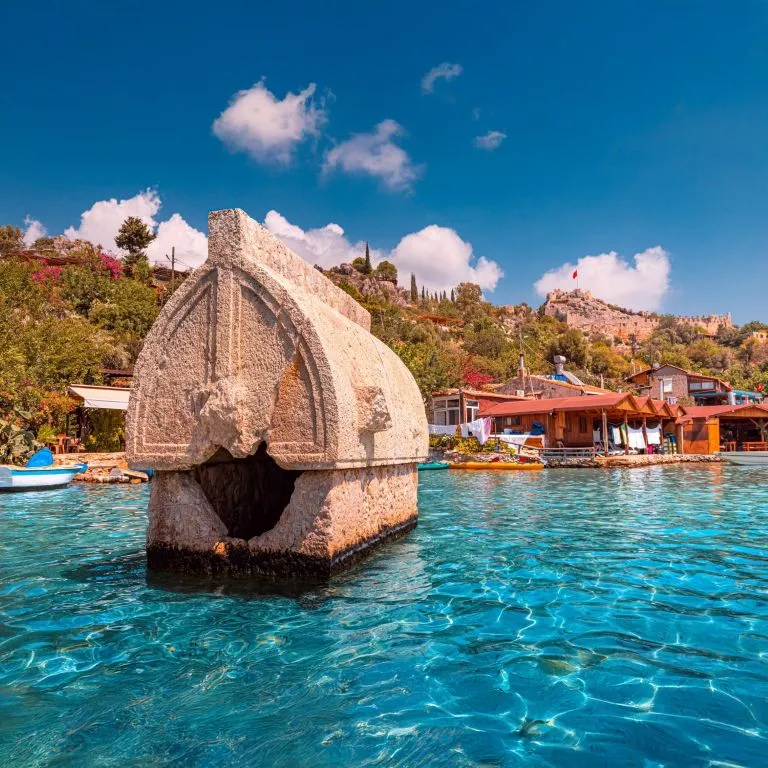 Ancient Lycian sarcophagus in the water at Kalekoy during the Demre to Kas and Back gulet cruise, with village houses and Simena Castle in the background