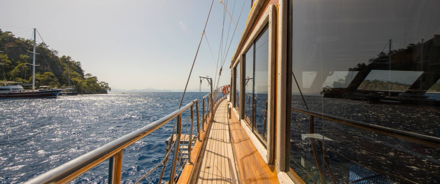 View along the wooden deck of a gulet yacht during a Sail Turkey blue cruise, with sparkling Mediterranean waters and forested coastline in the background