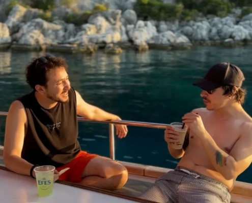 Two men enjoying drinks and relaxing on a gulet deck during a Kas to Kekova and Back gulet cruise in Turkey, with rocky green coastline in the background.