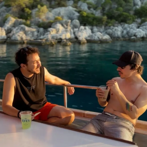 Two men enjoying drinks and relaxing on a gulet deck during a Kas to Kekova and Back gulet cruise in Turkey, with rocky green coastline in the background.